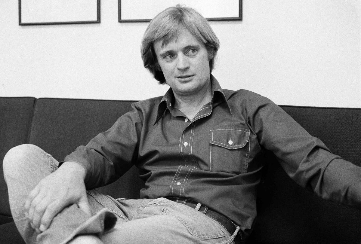 A black-and-white image of David McCallum lounging in a couch in a denim outfit