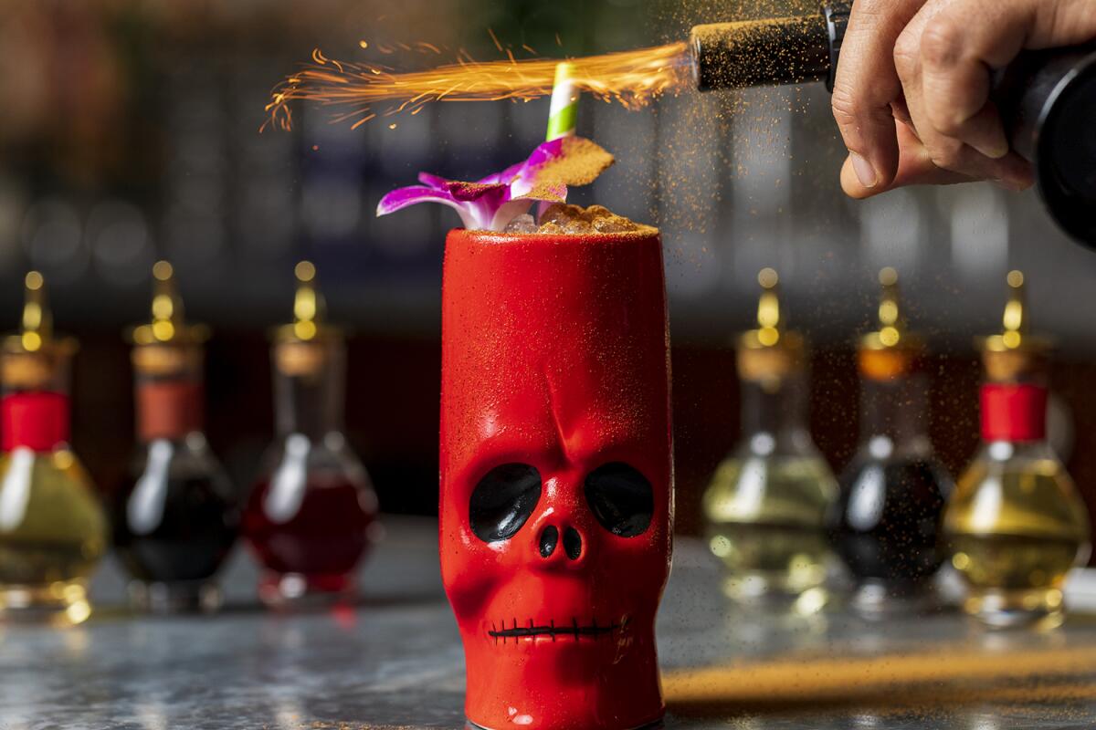 A person uses a small torch to flame the top of a cocktail served in a tall red tiki mug shaped like a skull