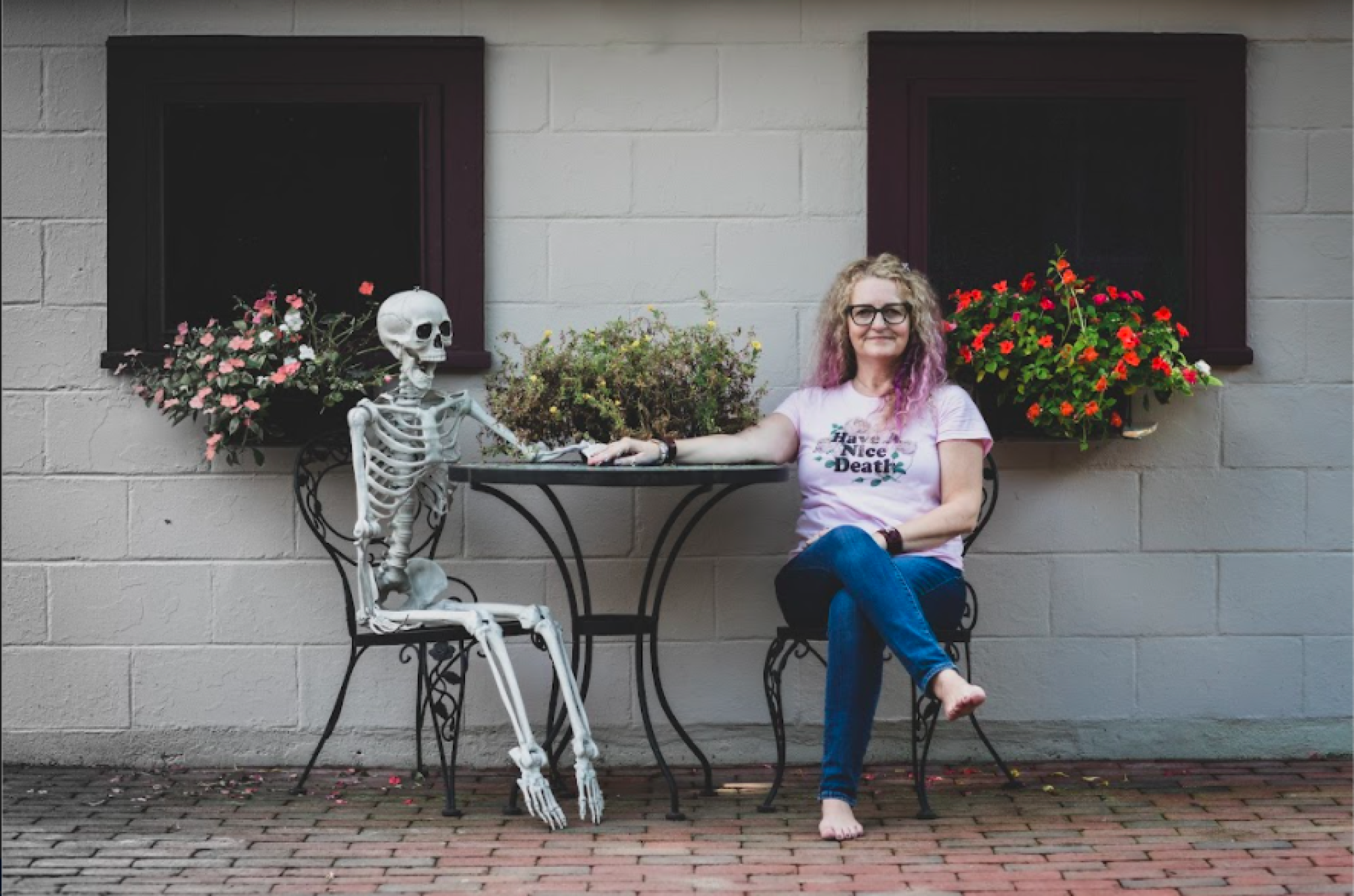 A fake skeleton and a woman sit at a table on a patio holding hands. 