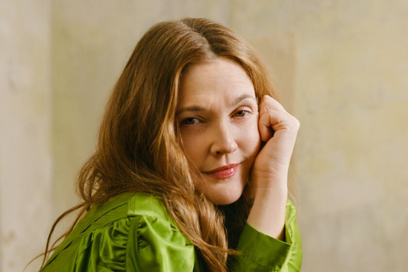 Drew Barrymore sits sideways in a shiny green outfit with arms crossed and one hand by her left cheek