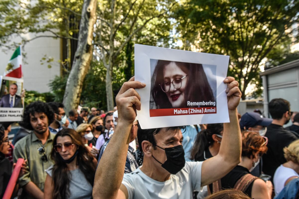 A crowd of protesters, one holding a photo of a woman with the words "Remembering Mahsa (Zhina) Amini."