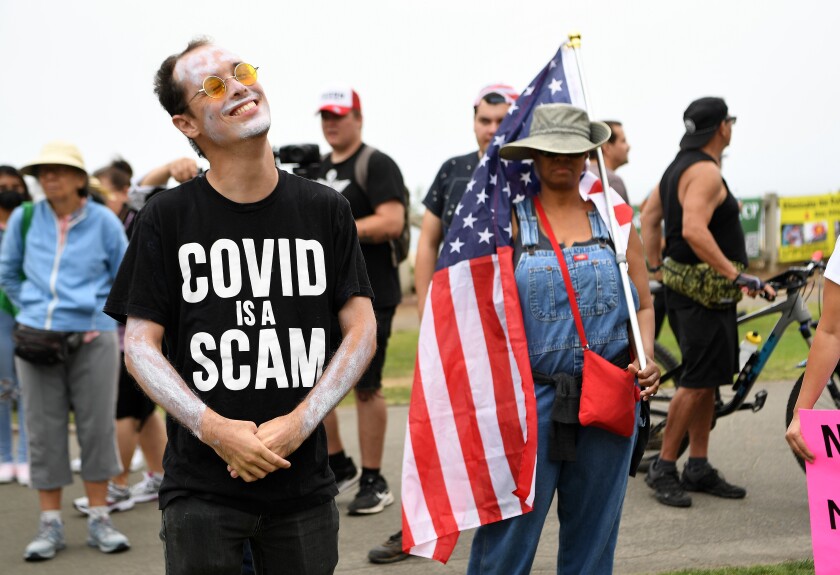 A protester in a T-shirt reading "COVID is a scam" stands next to a woman draped in an American flag.