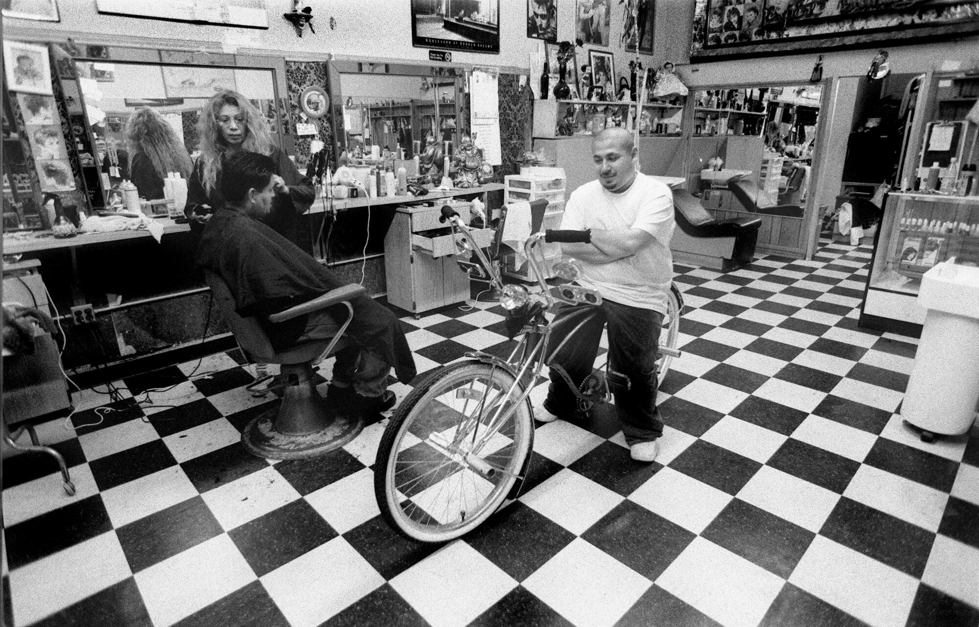 A man sits astride a bicycle in the middle of a salon as a woman cuts a customer's hair.