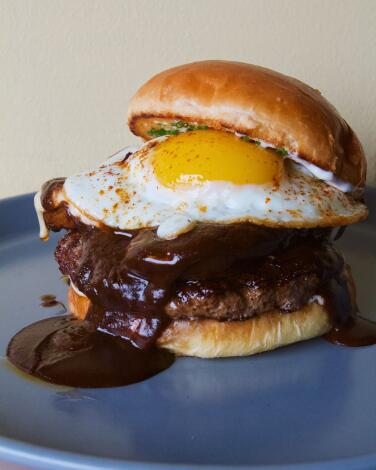 A vertical closeup of Macheen's mole burger topped with mole poblano, manchego, garlic aioli, mushrooms, chives and egg.