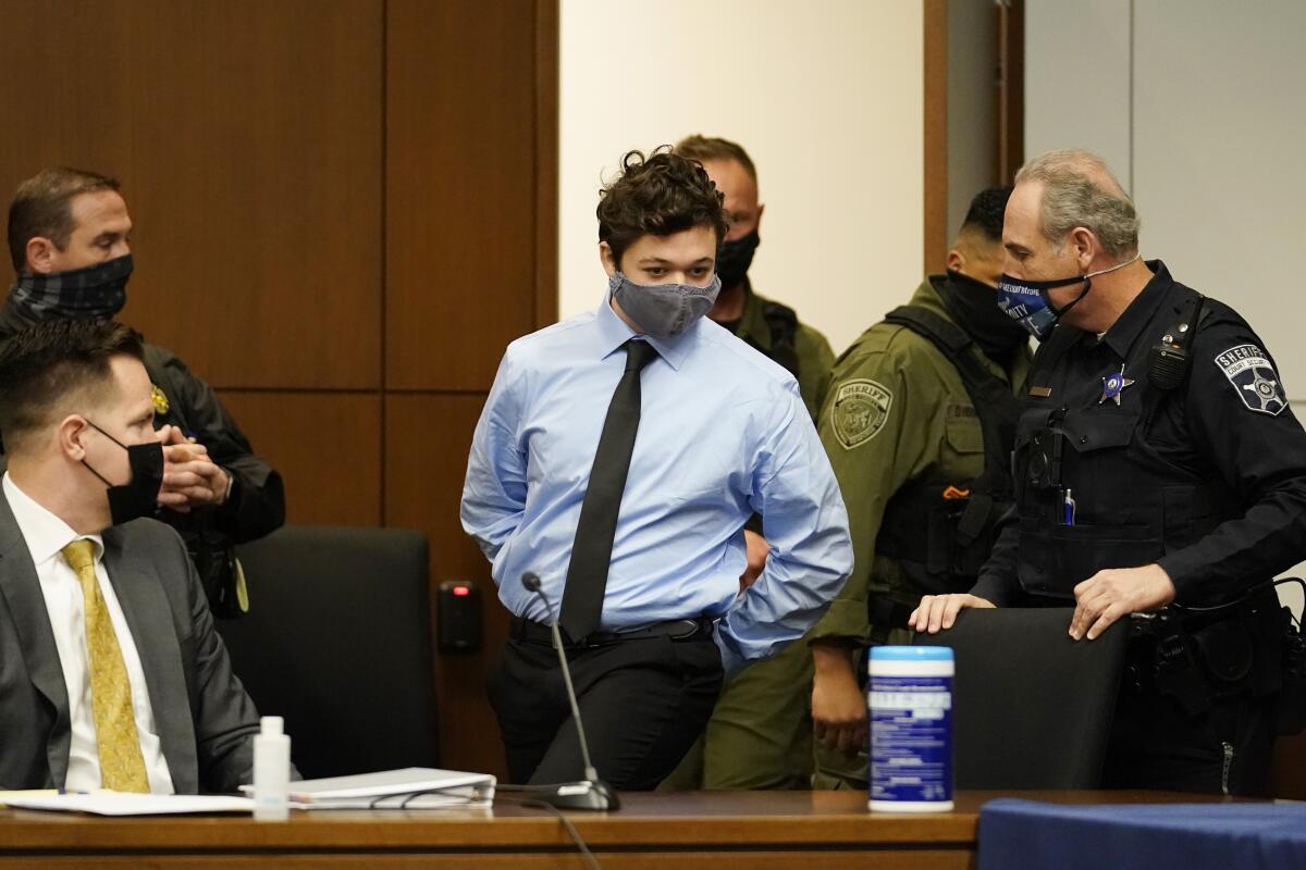 Teenager Kyle Rittenhouse, in collared shirt and tie, walks with his hands cuffed to a seat in court