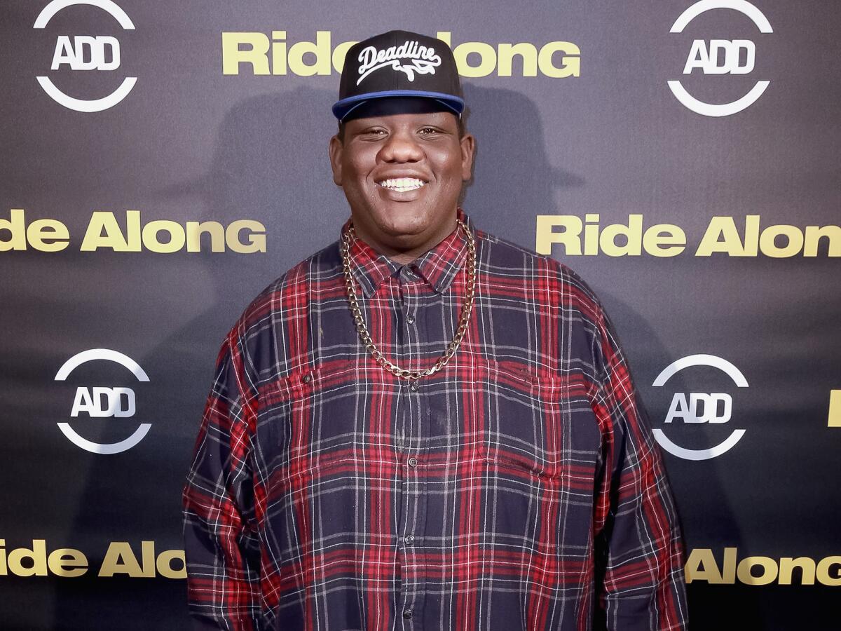 Teddy Ray stands in a baseball cap in front of a wall with logos