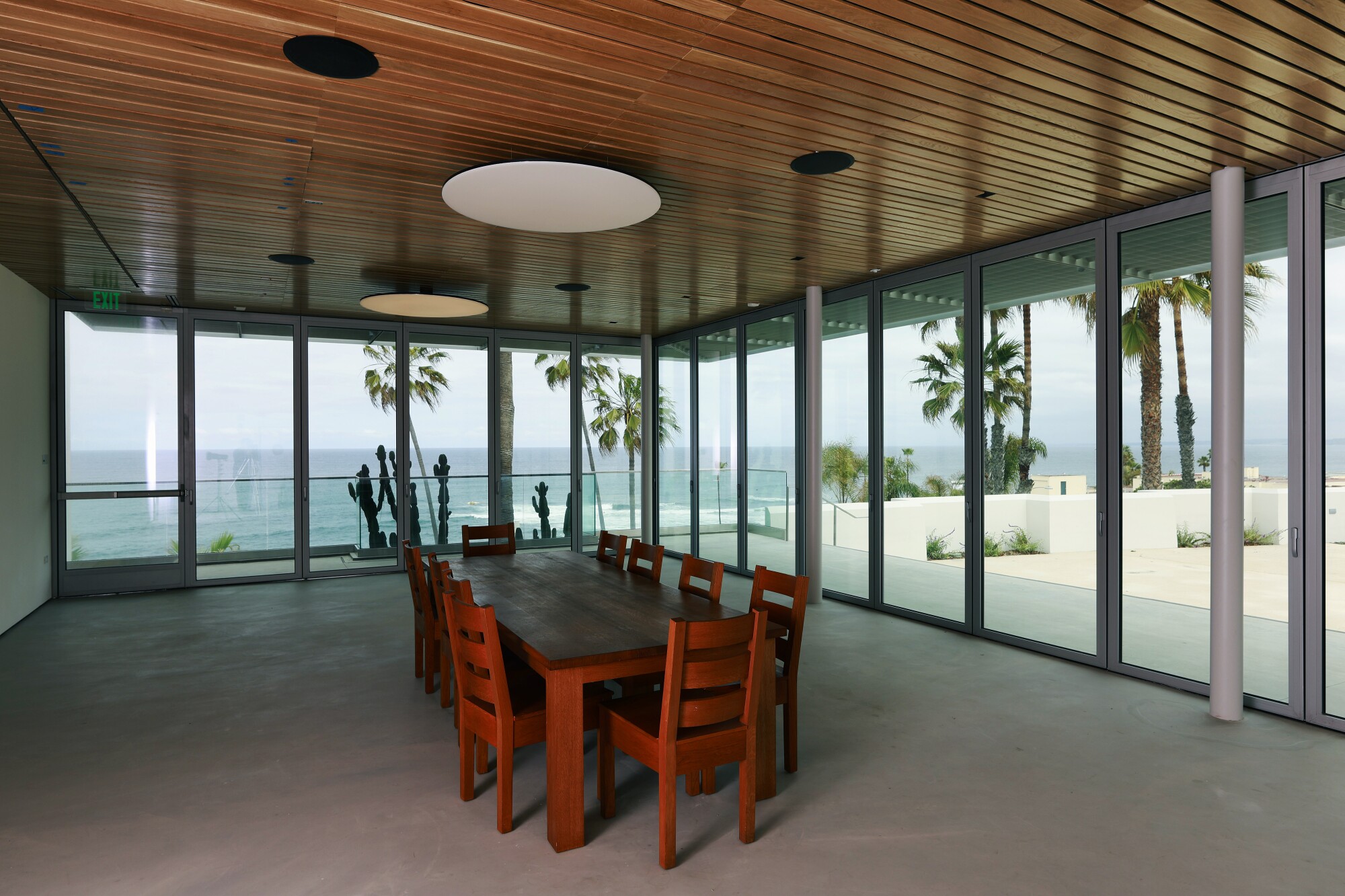 A red dining table in the middle of a room, with views of palm trees and the ocean seen through  glass walls