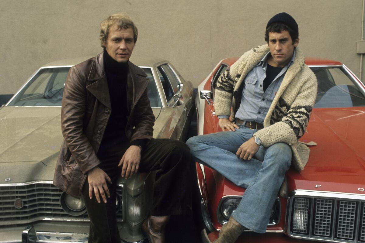 Two men sits side by side on the hoods of cars