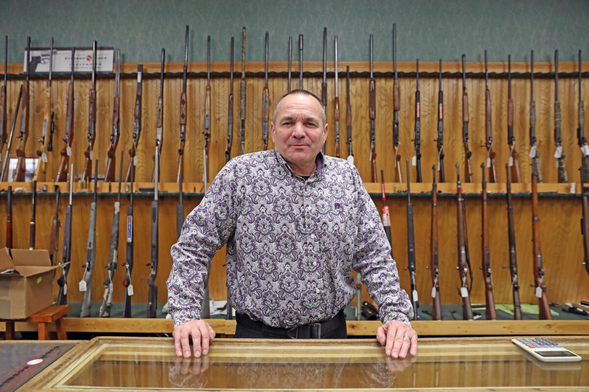 Patrick Jones stands at the counter of a gun shop, with rifles lined up behind him