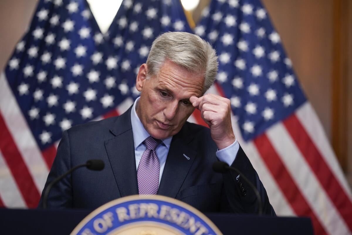 A man scratching his brow as he speaks in front of U.S. flags 