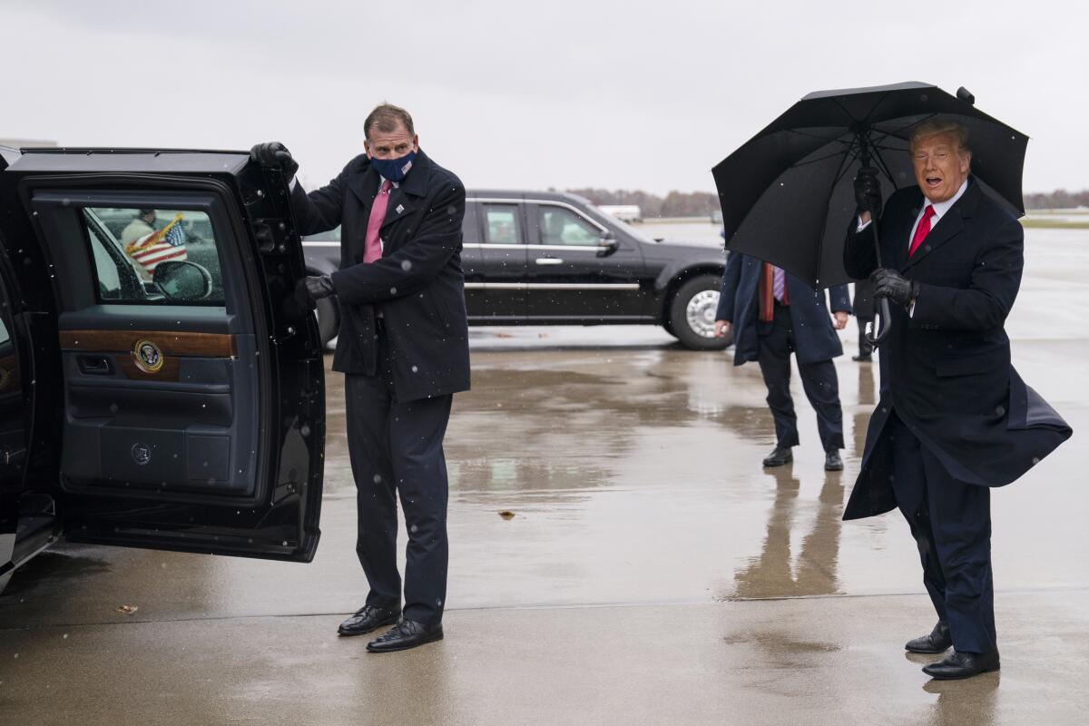 A man in a mask holds open the door of Trump's limo as the president holds an umbrella against the wind and rain.
