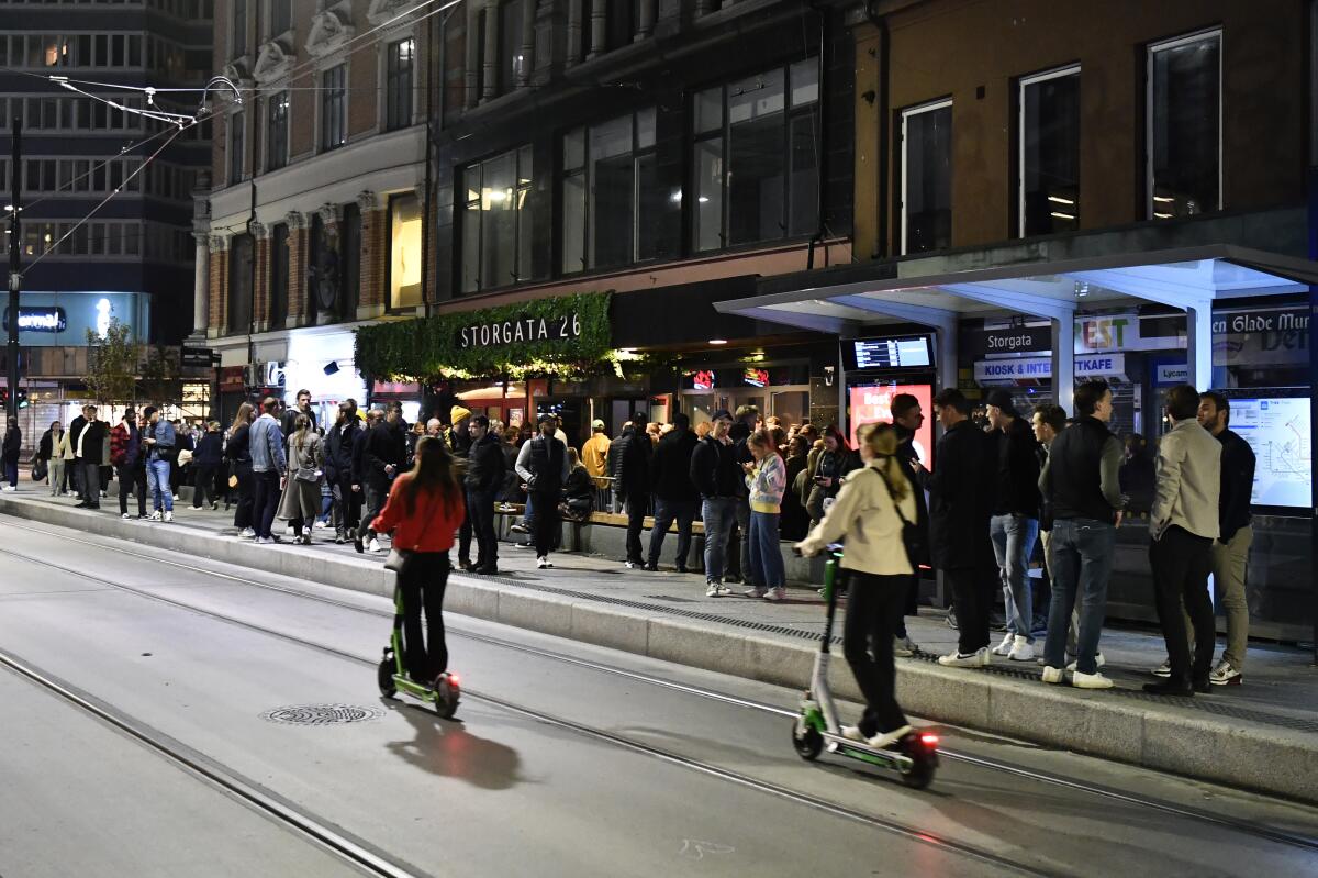 People stand on the sidewalk and ride scooters in Oslo.