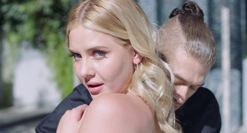 A blonde woman looks at the camera as a man hugs her in the movie “Pleasure.”