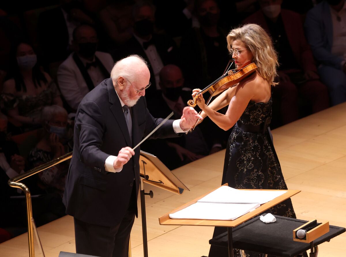 A man conducts an orchestra standing next to a woman in a black dress playing the violin.