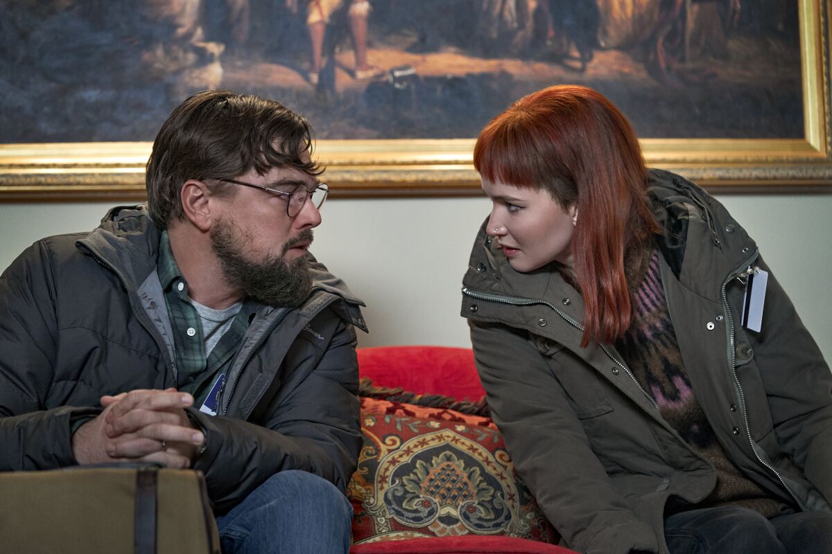 A bearded man and a red-haired woman lean together to talk on a sofa.