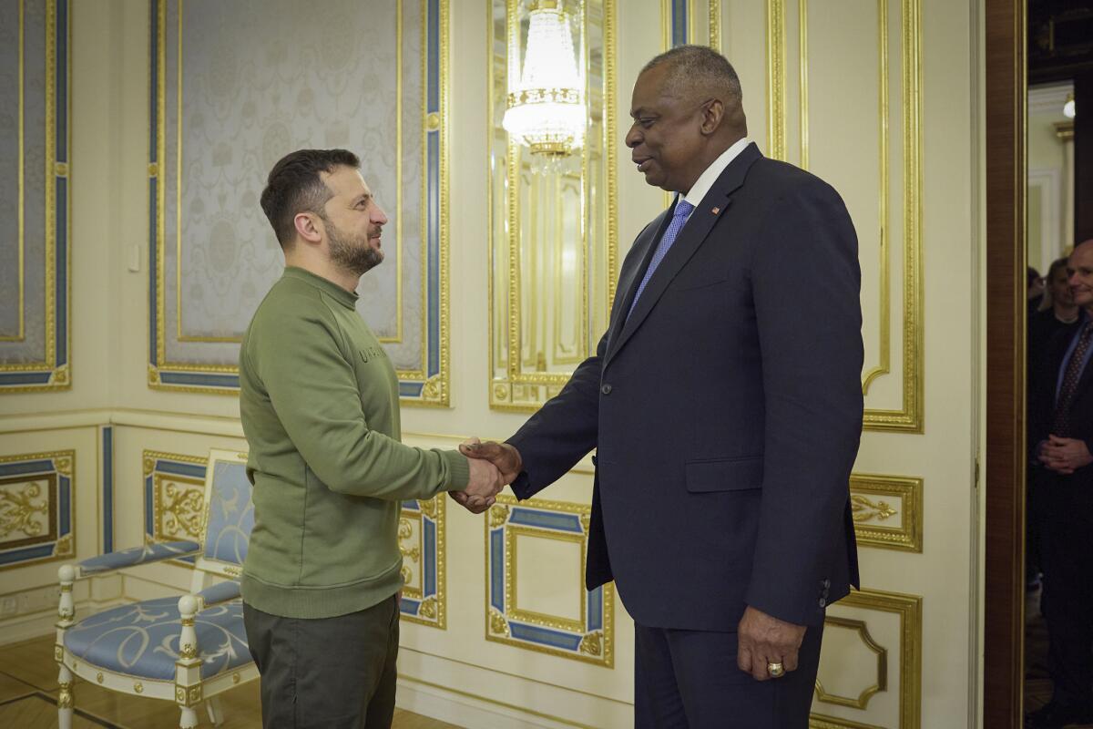 A man with dark hair and beard, in a green shirt, left, and a taller man in dark suit and tie shake hands in an ornate room