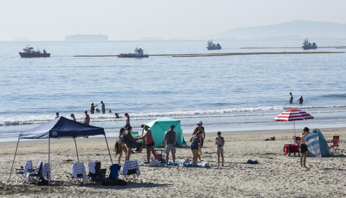 People on the beach and in the ocean as tugboats pull oil booms offshore