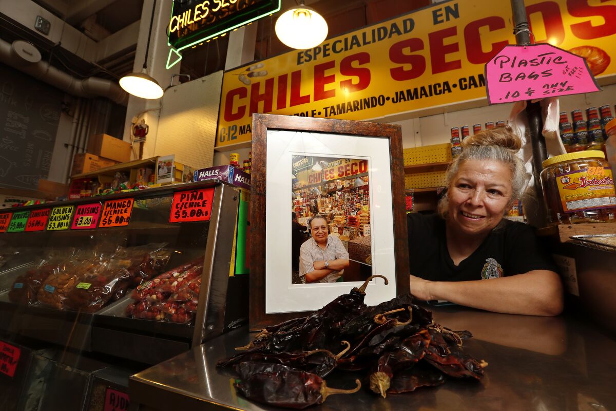 A smiling woman at a food counter holds up a framed photo of a man with his arms crossed 