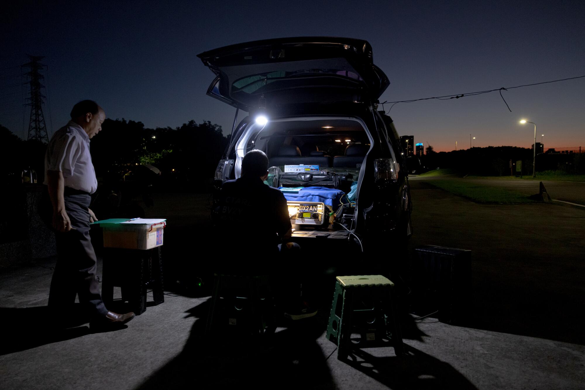 A man, left, stands near another man who is seated in front of the open trunk of a van, with a bright bulb providing light