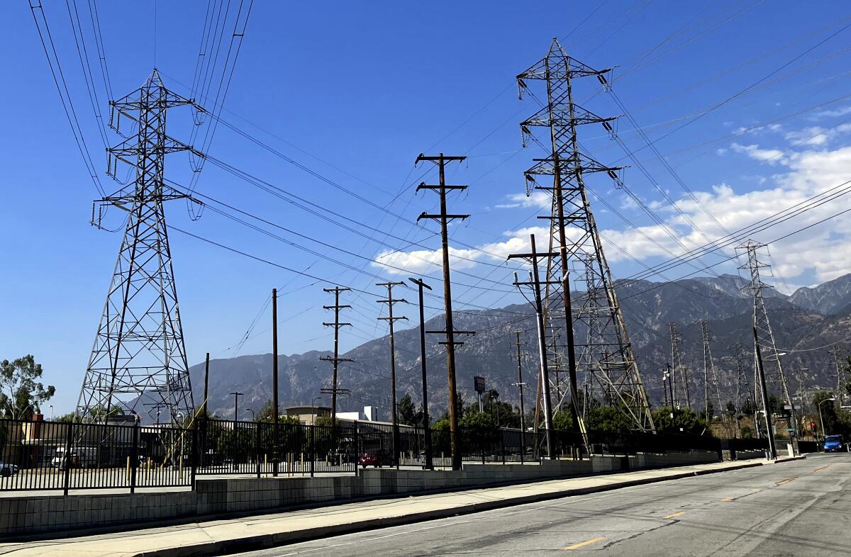 Electrical grid towers are seen in front of hills under a blue sky with puffy white clouds