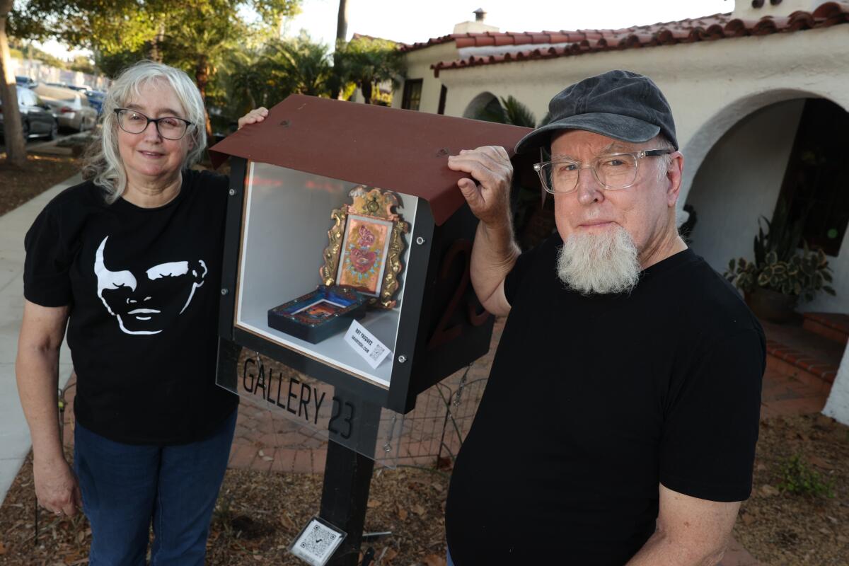 Dave and Carol Clark stand beside a micro art gallery, which looks a bit like an open, oversized residential mailbox.