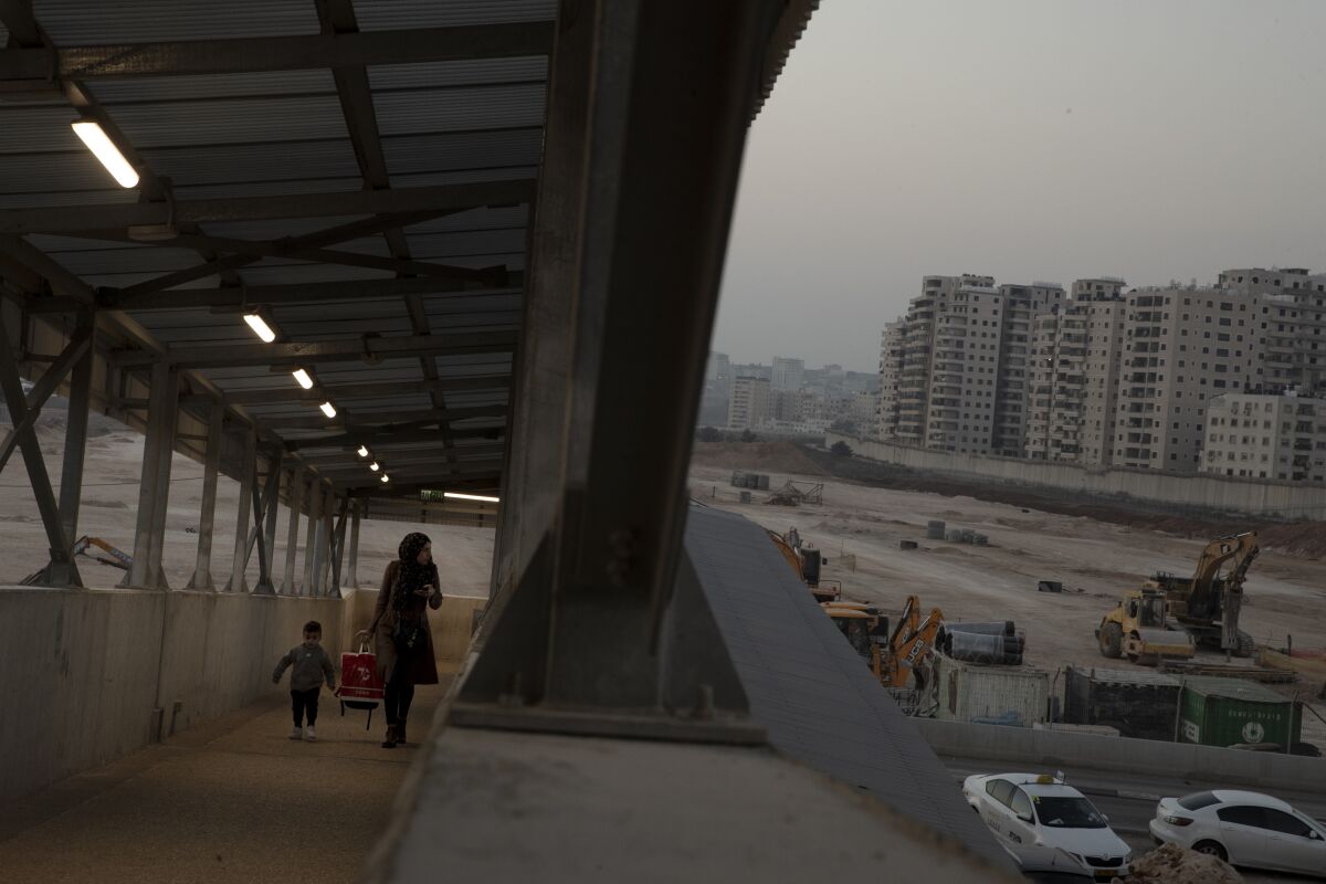 A woman and a child walk up a ramp. To the right are construction vehicles with buildings in the background.
