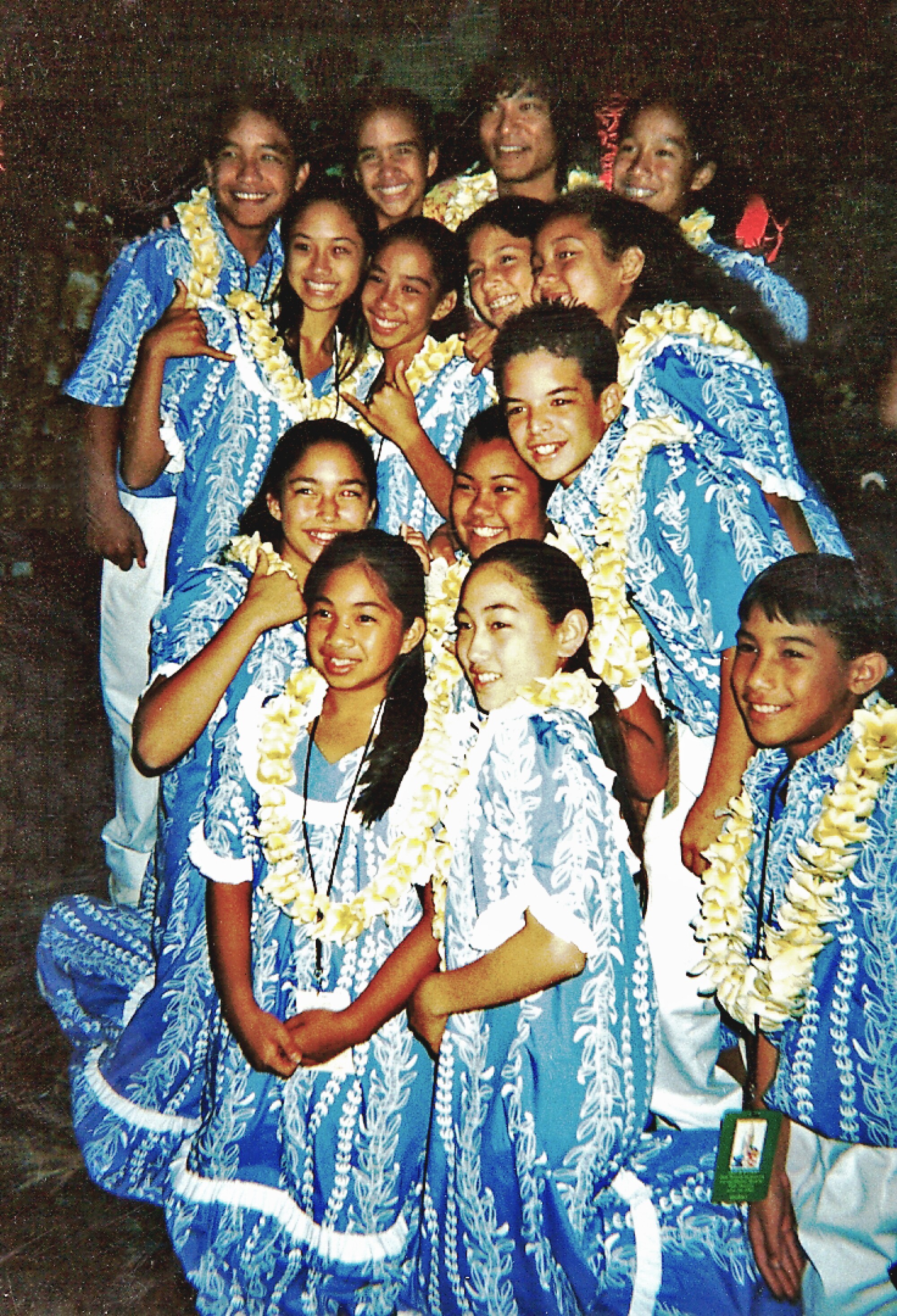 A group of children in matching, ruffled blue outfits and yellow leis posing with a man