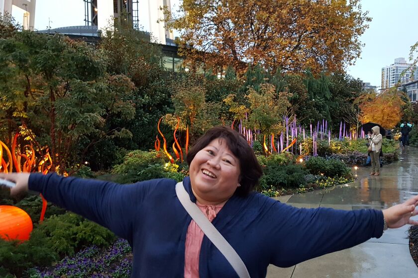 Sally Fontanilla in Seattle with a big smile and her arms spread wide. Behind her, autumn foliage lining a walkway.