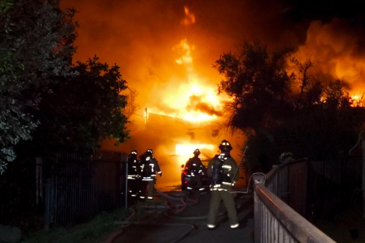 Firefighters stand in front of a burning home in the predawn dark.