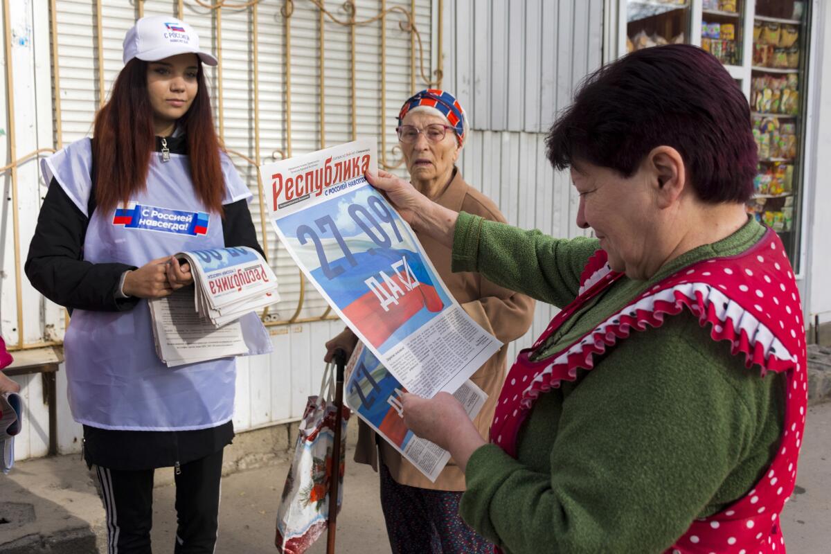A woman in a red apron, right, holds up a newspaper to read as two other women, one holding newspapers over one arm, look on
