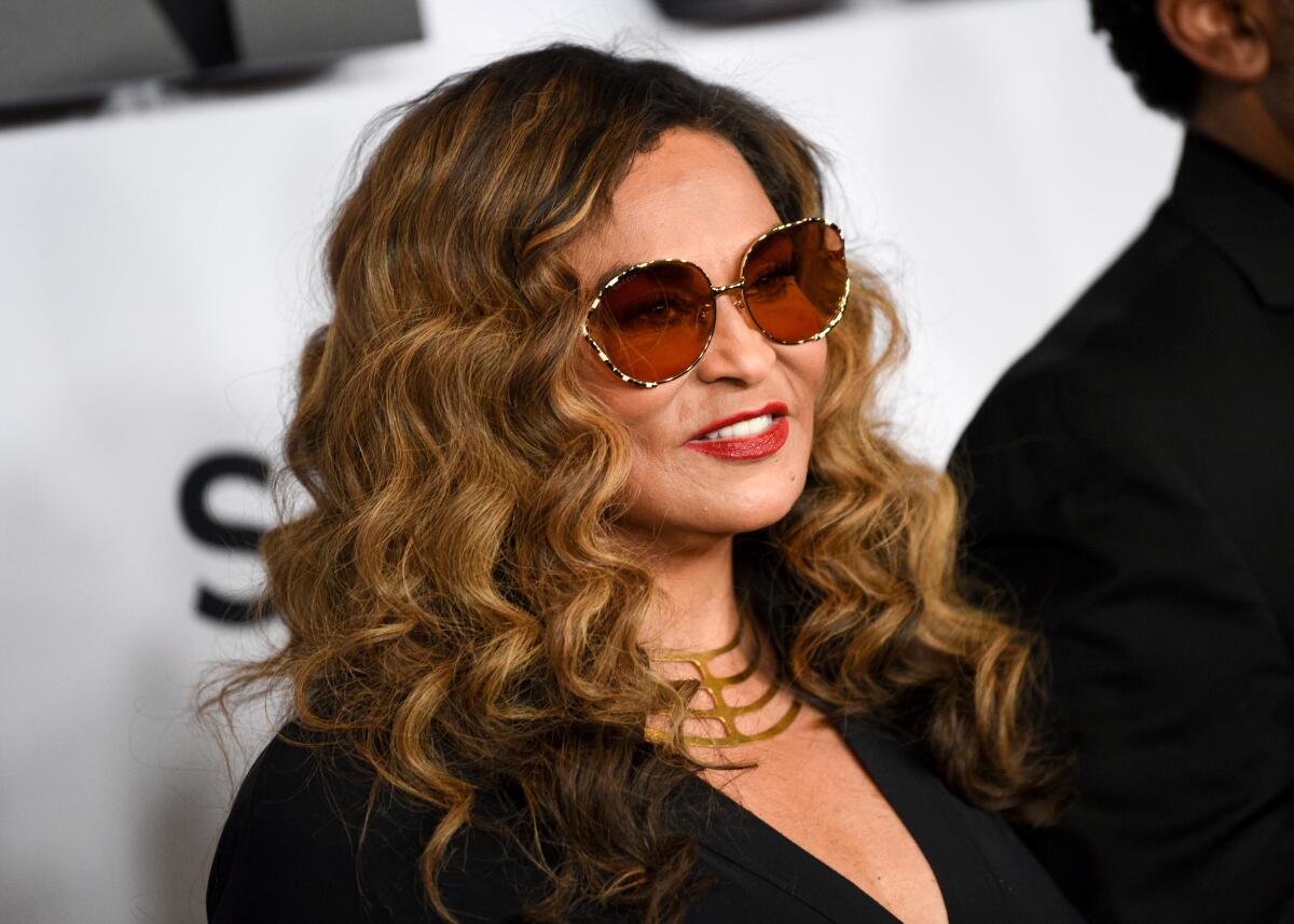 Tina Knowles in a black dress, big sunglasses and a gold necklace smiling and posing against a white background