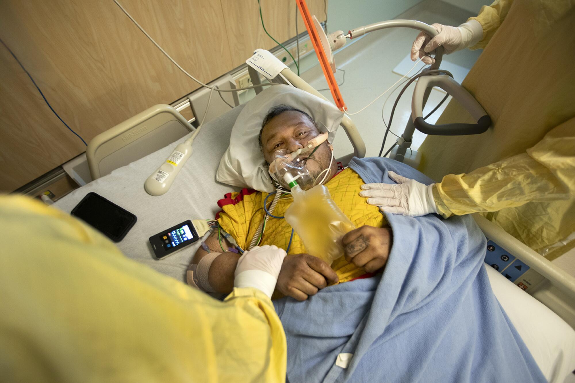 Two healthcare providers in protective gear place their hands on a man in a hospital bed 