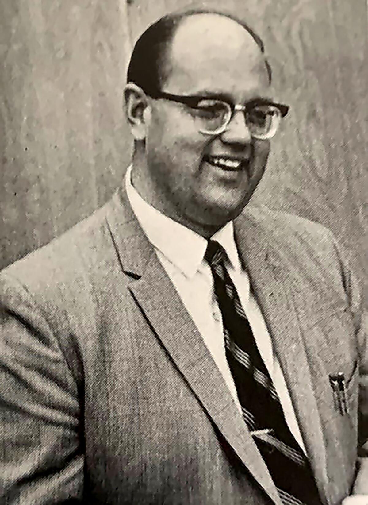 A smiling man in suit and tie and wearing glasses, with pens in his jacket pocket 