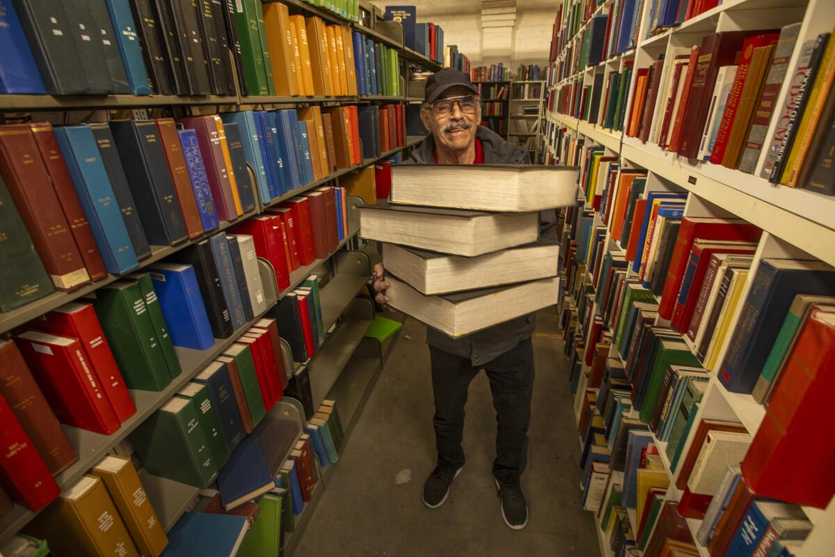 A man holds a stack of large books between two long bookshelves.