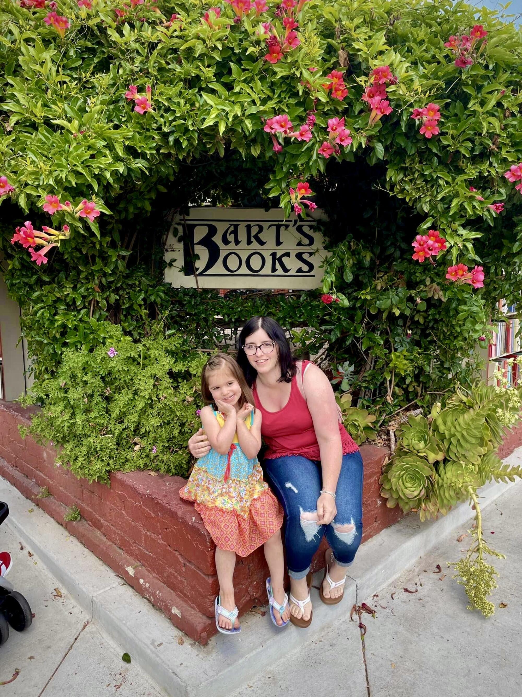 A mother and daughter sit in front of the Bart's Books sign, surrounded by flowers and greenery.