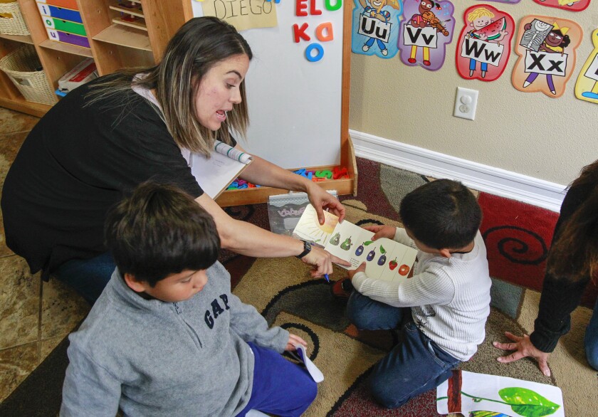 A woman in a classroom reads a picture book with a little boy as another boy plays