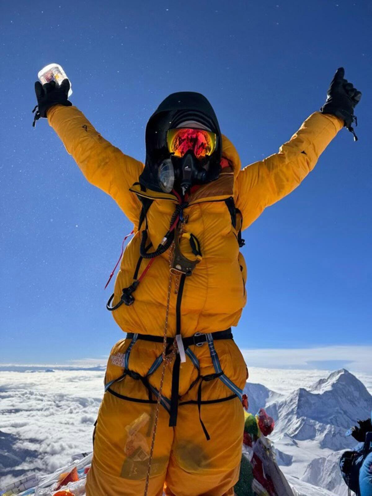 A man in yellow snowsuit stands with arms raised on the summit of Mt. Everest.