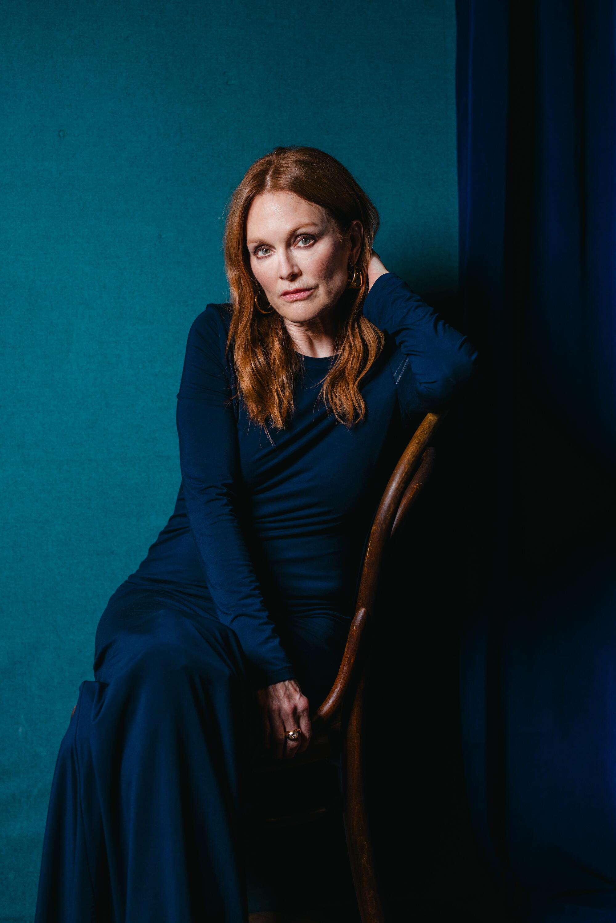 Julianne Moore sits on a wooden chair against a blue backdrop and curtain for a portrait.