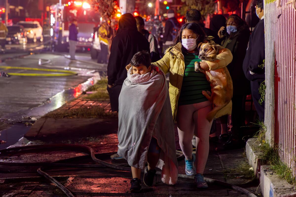 A woman in a mask carries a dog and walks next to a boy wrapped in a blanket with fire trucks in the background