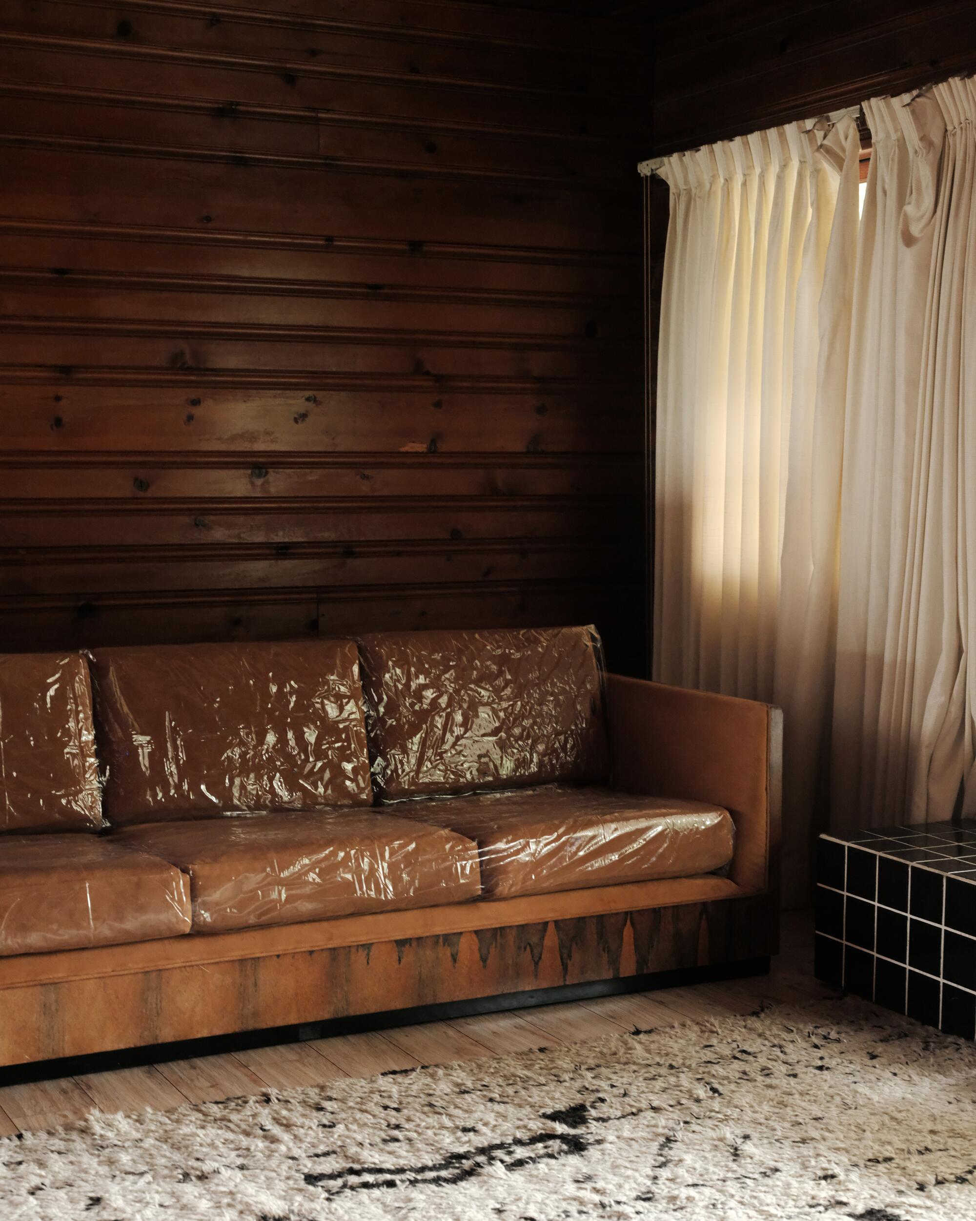 A photo of a plastic-covered brown couch, a wooden wall and white curtains in the background.