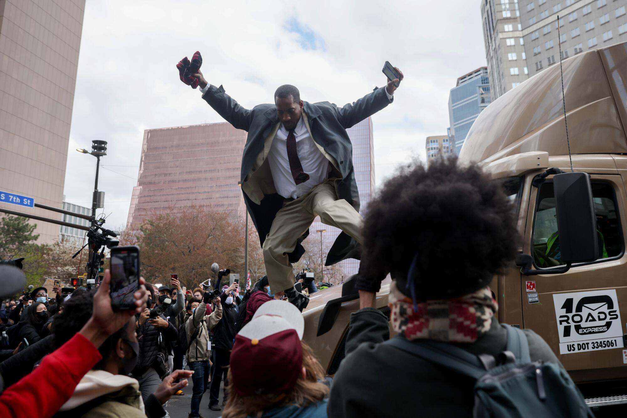 A man in a suit and tie jumps down from the hood of a truck 