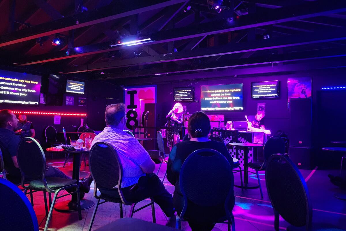 People sit in a colorfully lit room watching a karaoke singer, with screens showing lyrics around the room.