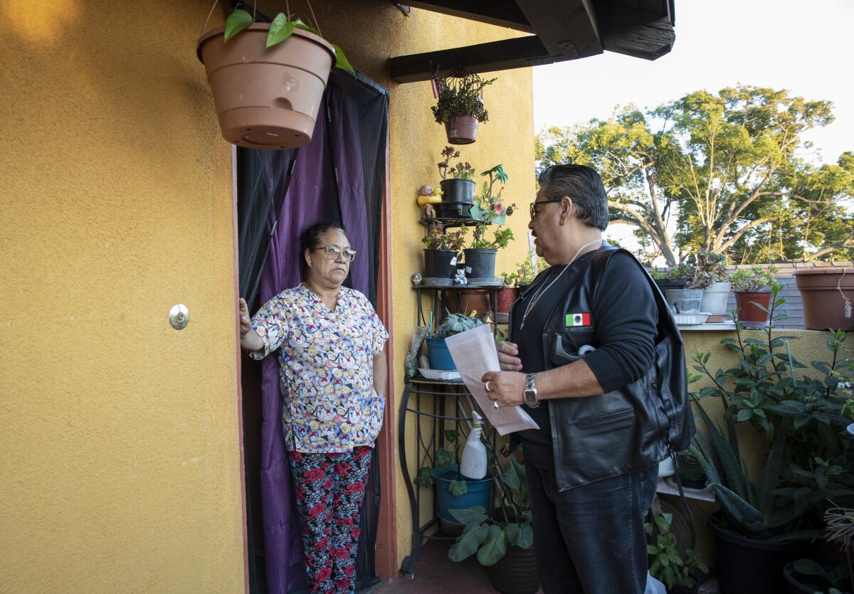 A woman stands at the door of a modest home, speaking with a man in a leather vest. 