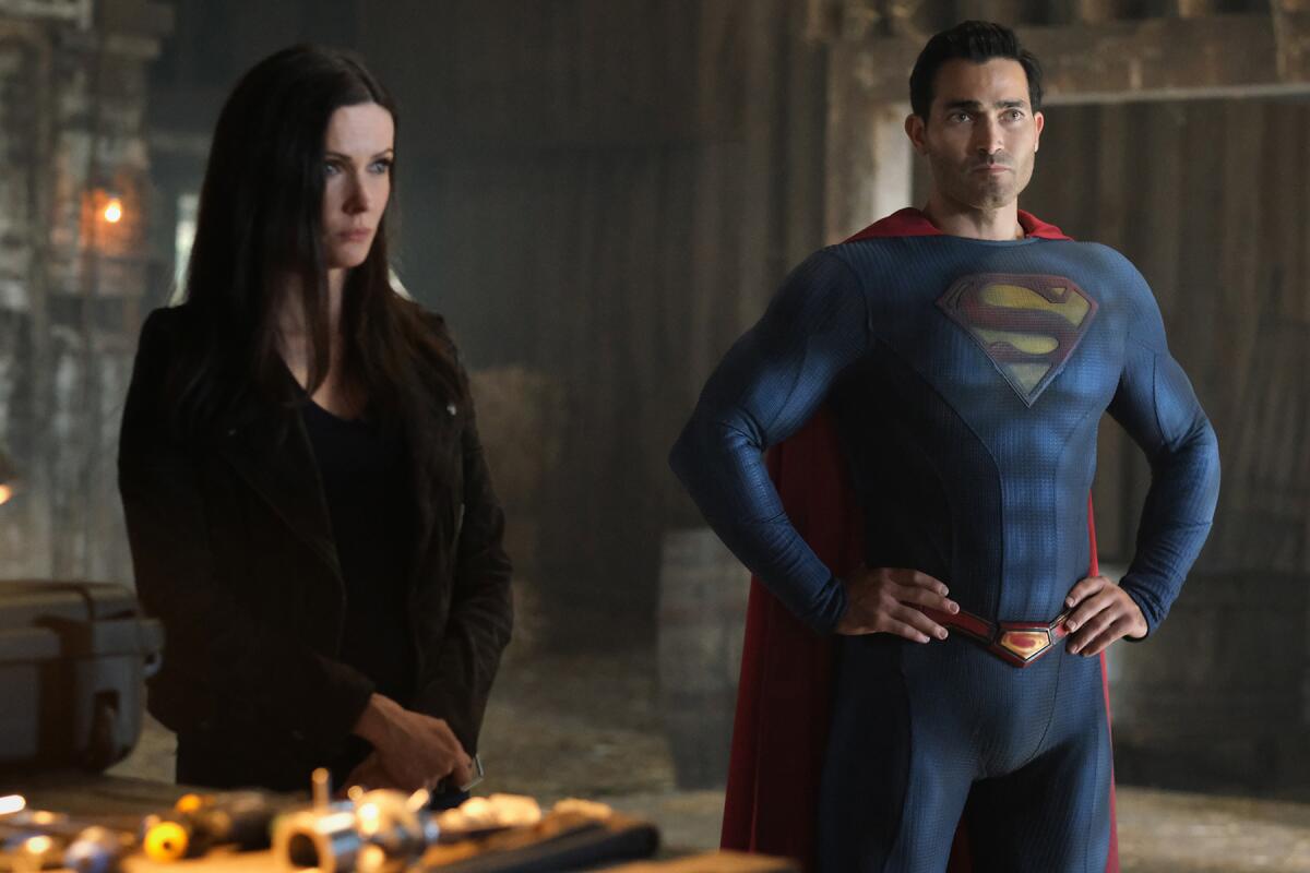 A woman and a man in a blue fitted suit with an S emblem on his chest and a red cape stand while looking forward.