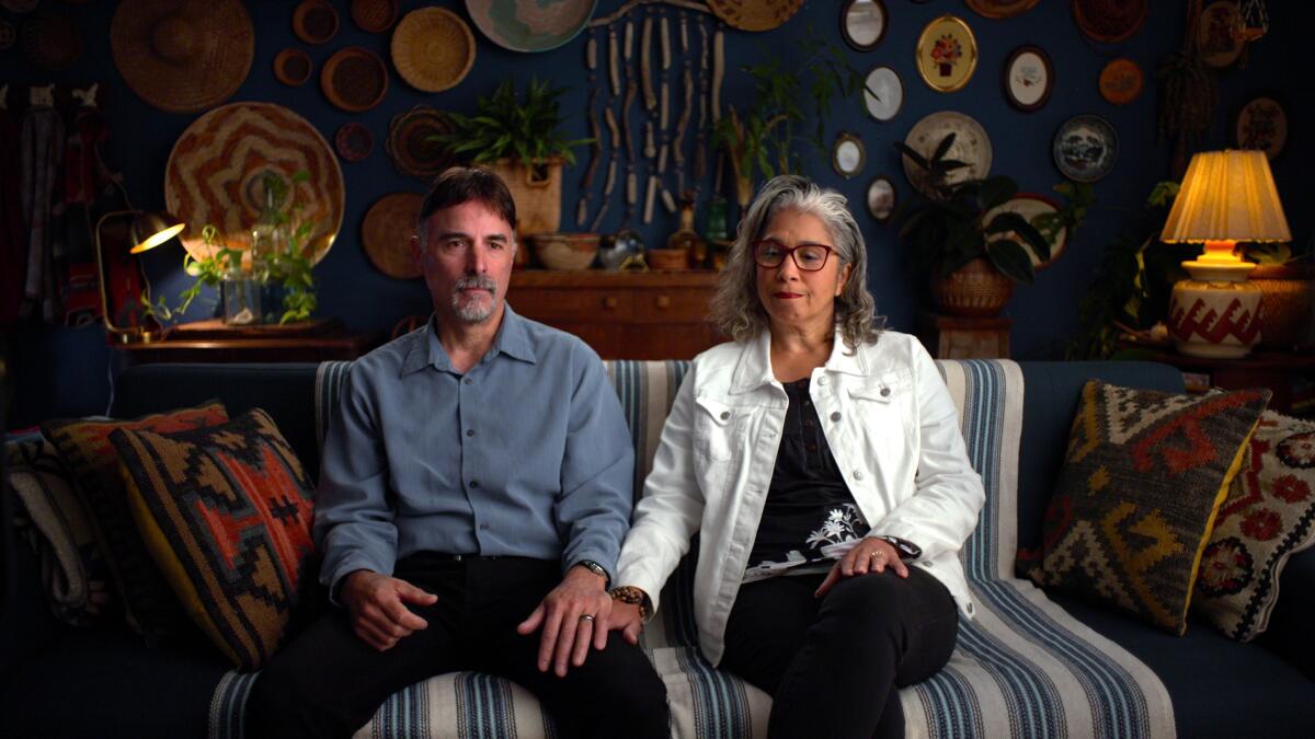 Lawrence and Migdalia Raiano sit on a couch in a busy room, his left hand over her right as she rests it on his leg