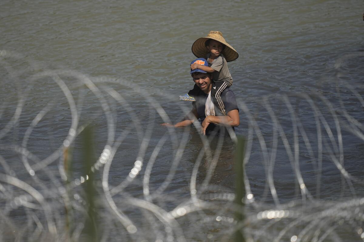 A man with a child on his shoulders stands in a river, behind concertina wire. 