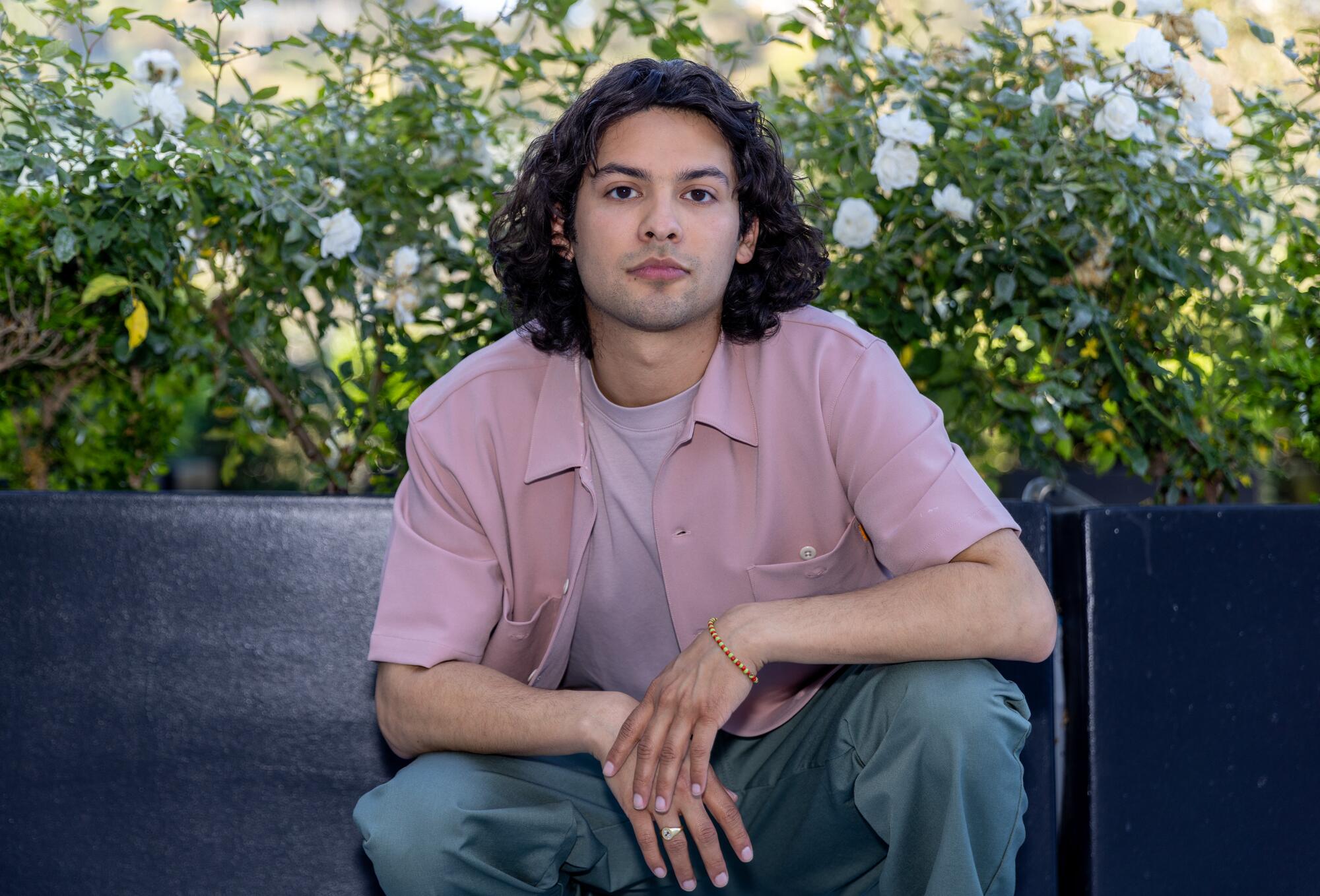 Xolo Maridueña poses in front of a rose bush. He's wearing a red button-up shirt and jeans.