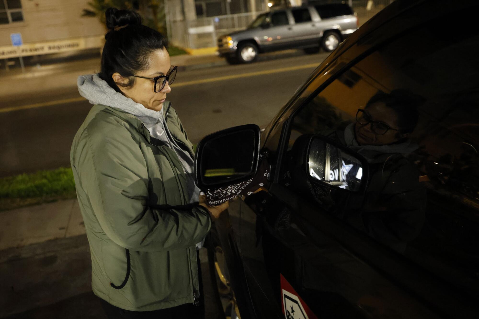 A woman in a jacket ties a bandana to a vehicle's mirror.
