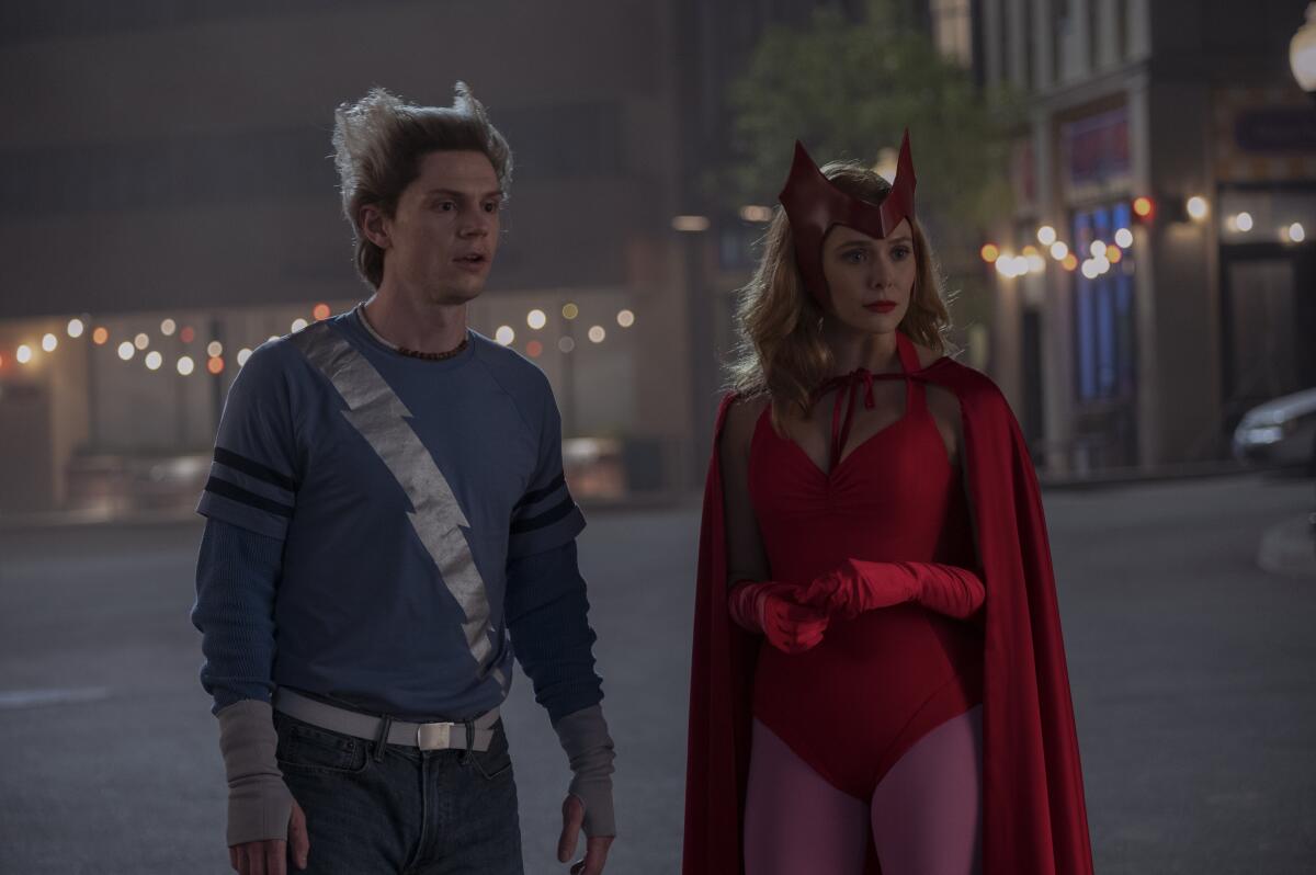 Evan Peters in a blue top with a silver lightning bolt and Elizabeth Olsen in a red leotard, gloves and cape in a street.