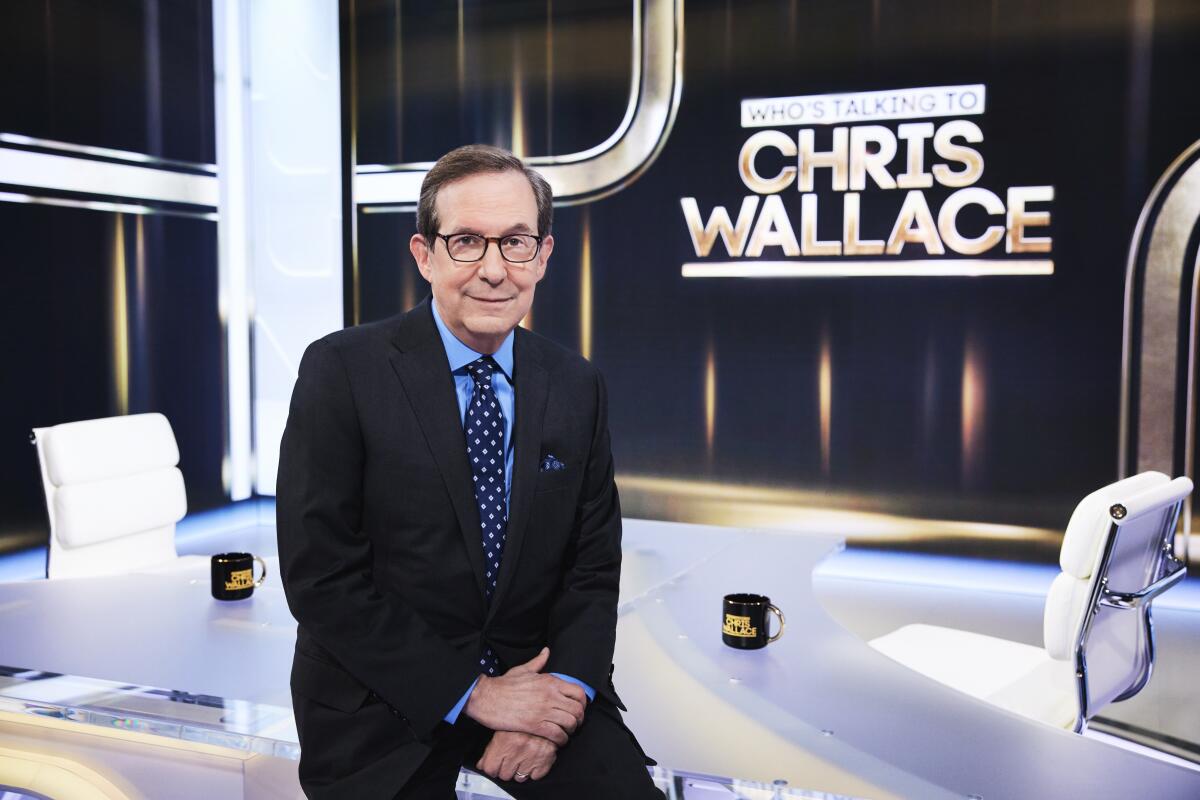 A man in a suit and glasses sits on a TV set in front of a sign that says "Who's Talking to Chris Wallace"
